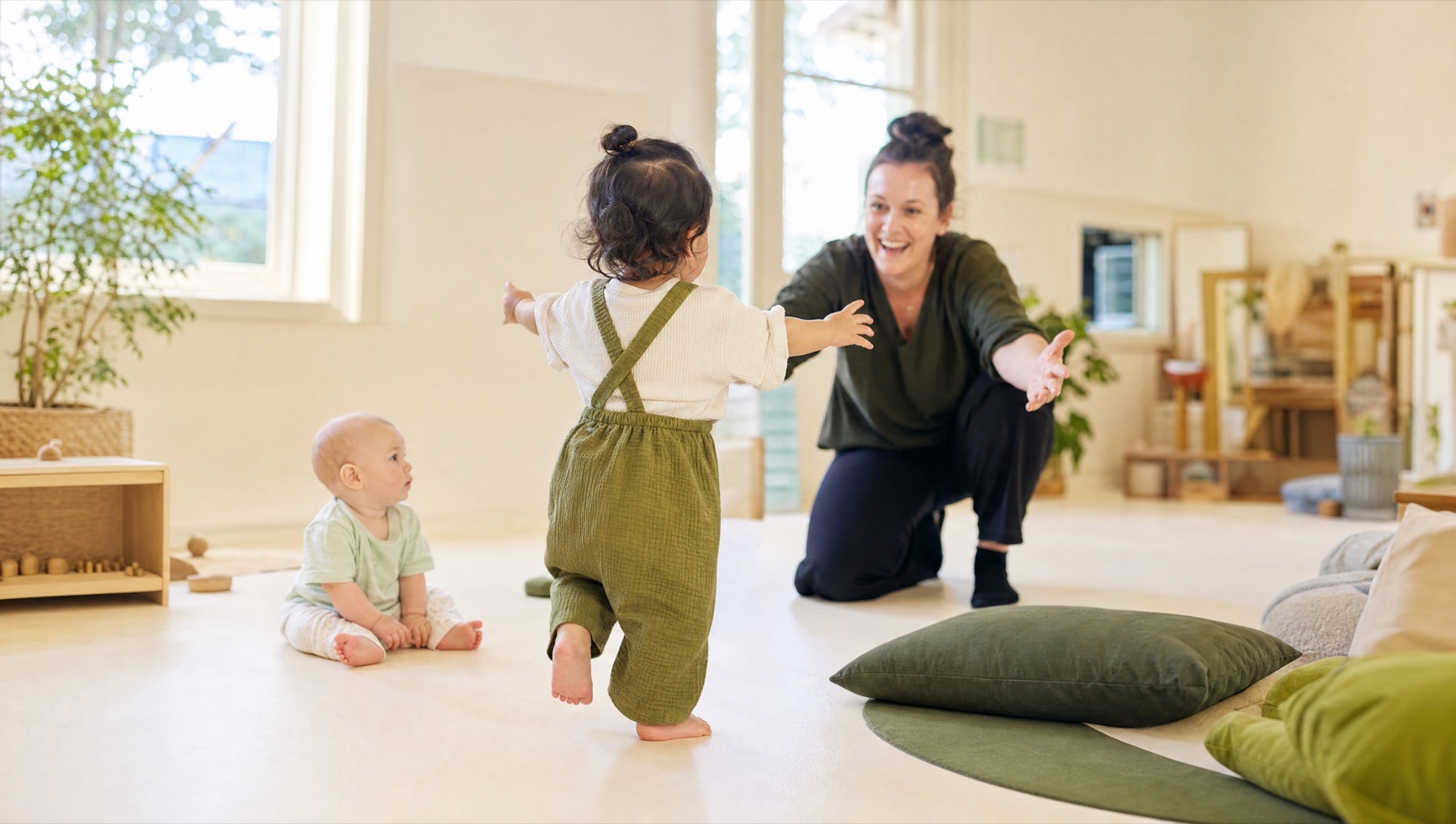 A toddler taking eager steps toward an educator's open arms in a bright early learning room